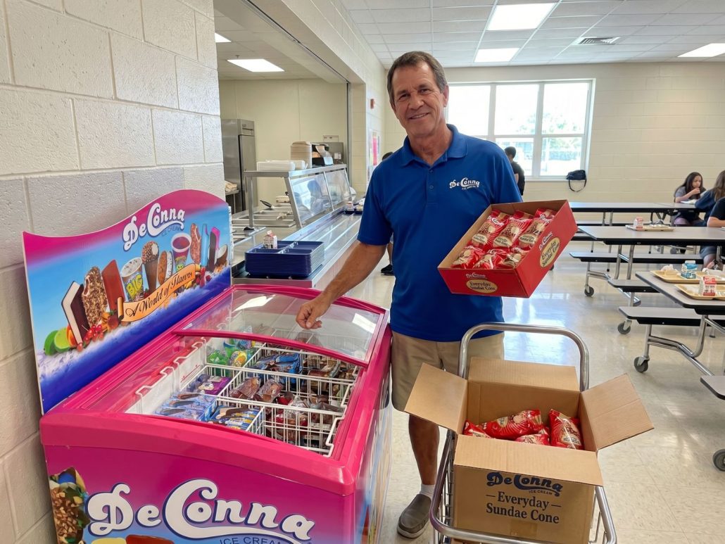 Smiling man in a blue polo stocks a pink DeConna ice cream chest in a school cafeteria.