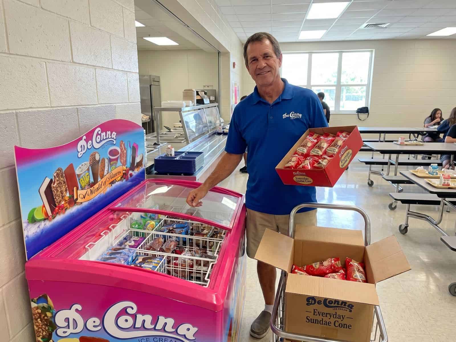 Smiling man in a blue polo stocks a pink DeConna ice cream chest in a school cafeteria.