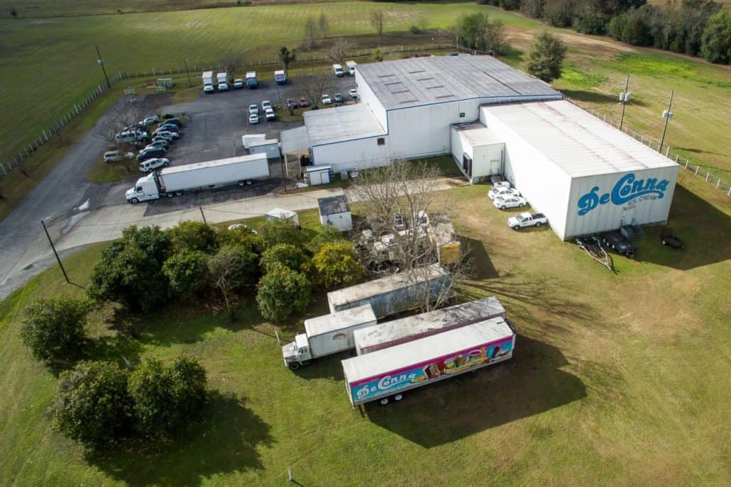 DeConna Distribution Center In Reddick - DeConna Ice Cream Aerial view of an ice cream factory complex with white buildings, loading docks, and multiple trucks parked in a lot surrounded by green fields.